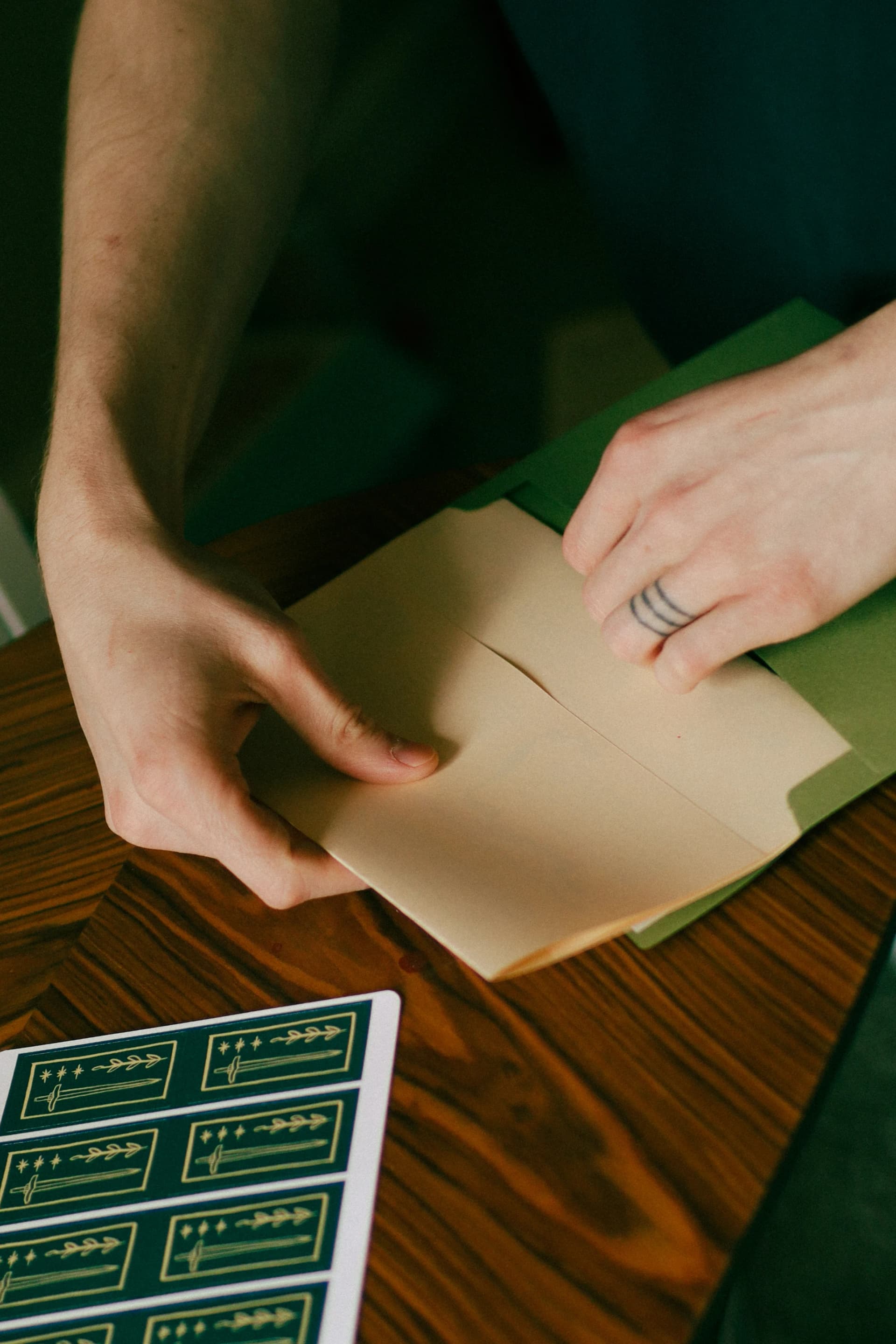 Hands packing a print into a green envelope beside gold-foil sword stickers on a wooden table