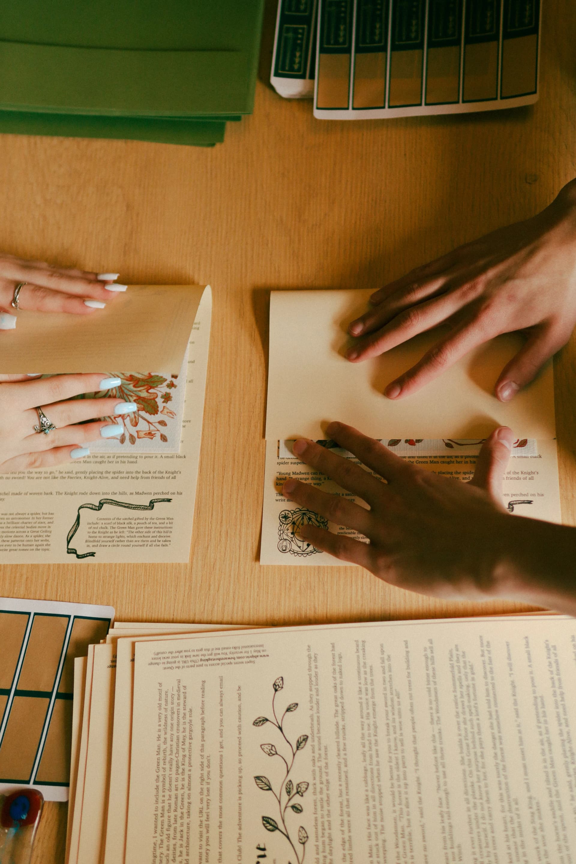 Two pairs of hands folding cream story letters with black ink illustrations on a wooden table