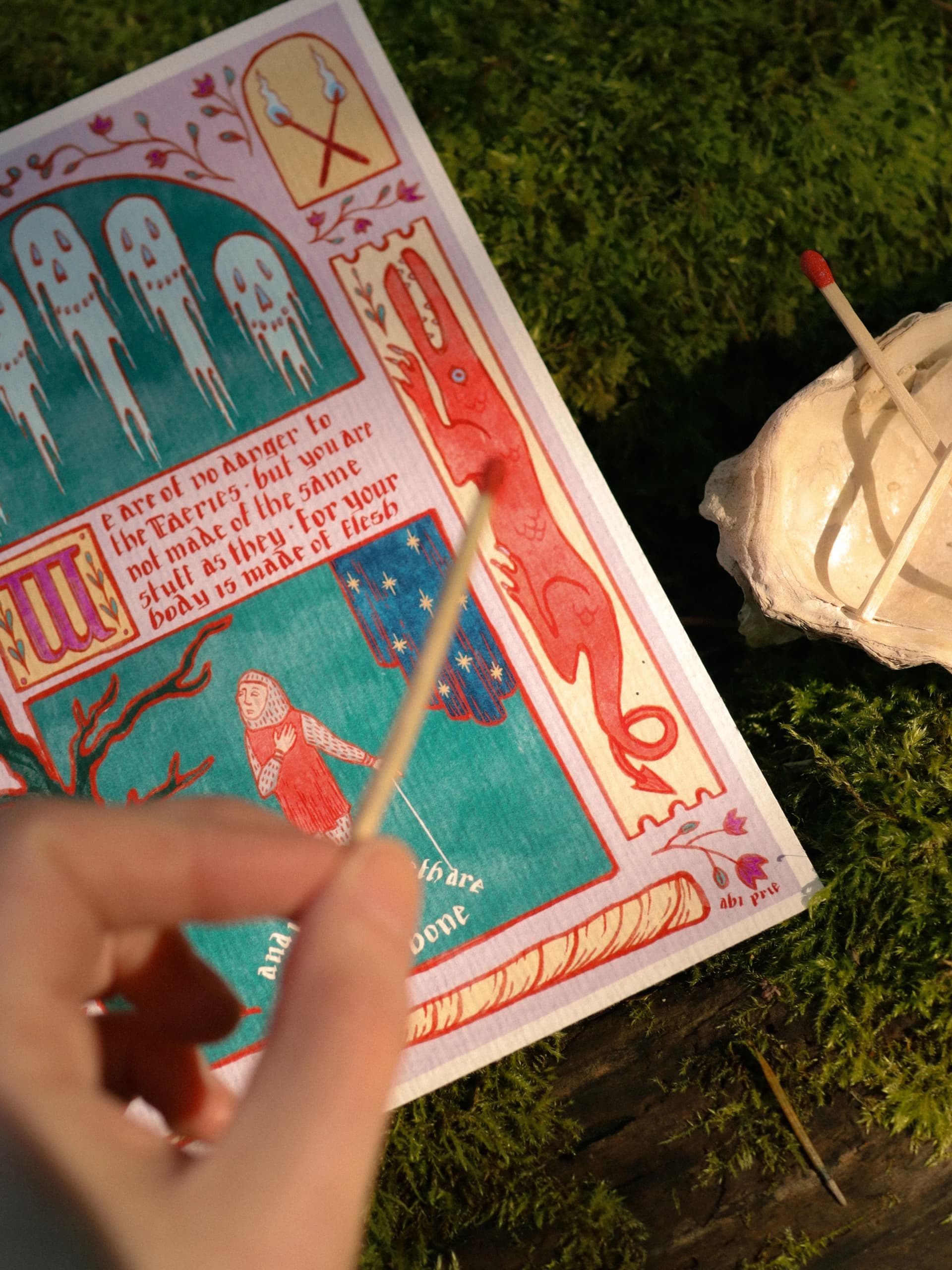 Illuminated manuscript-style Quest print on moss, with a hand holding a lit match toward the page