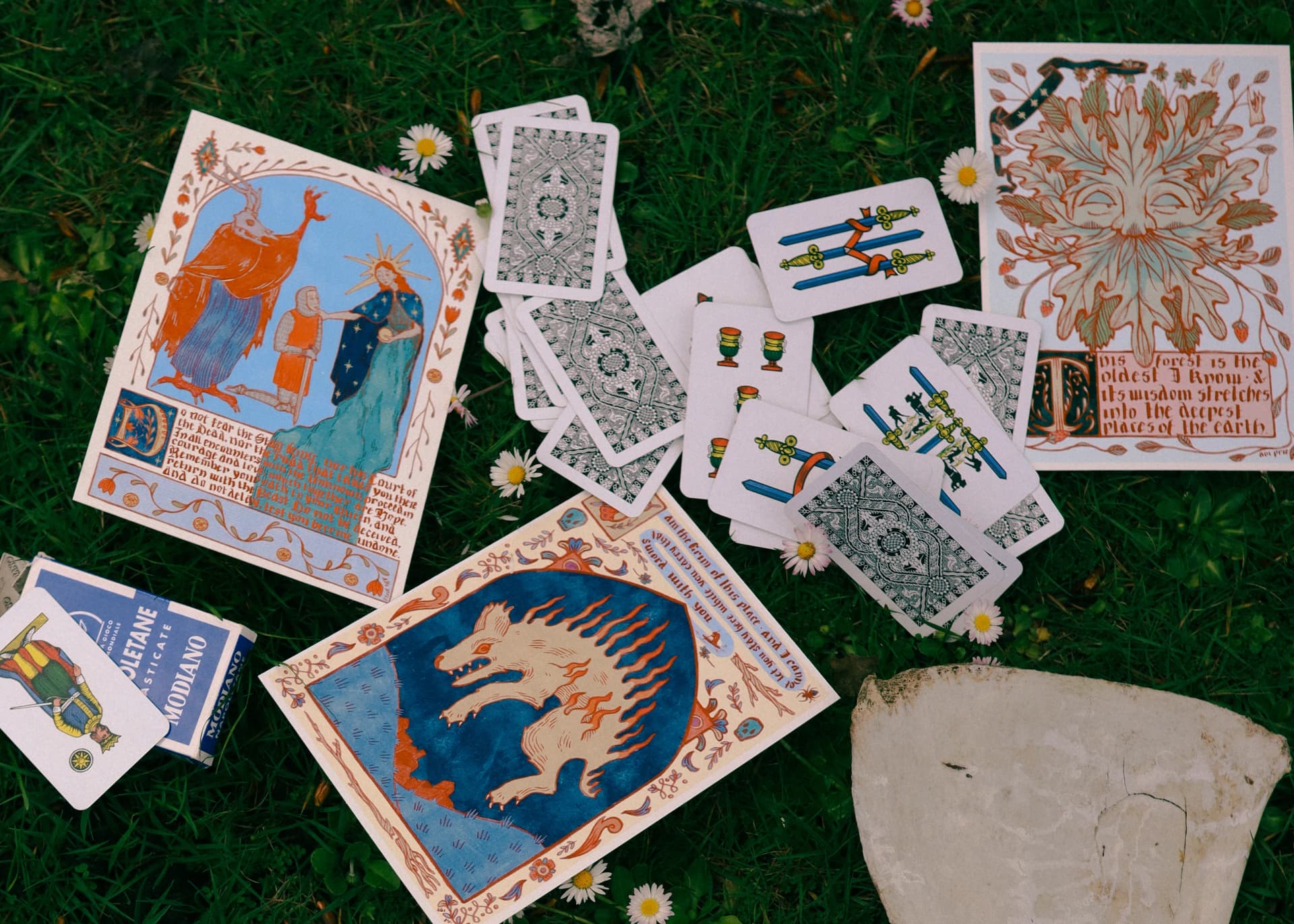 Folkloric Quest prints and playing cards arranged on grass among small white daisies
