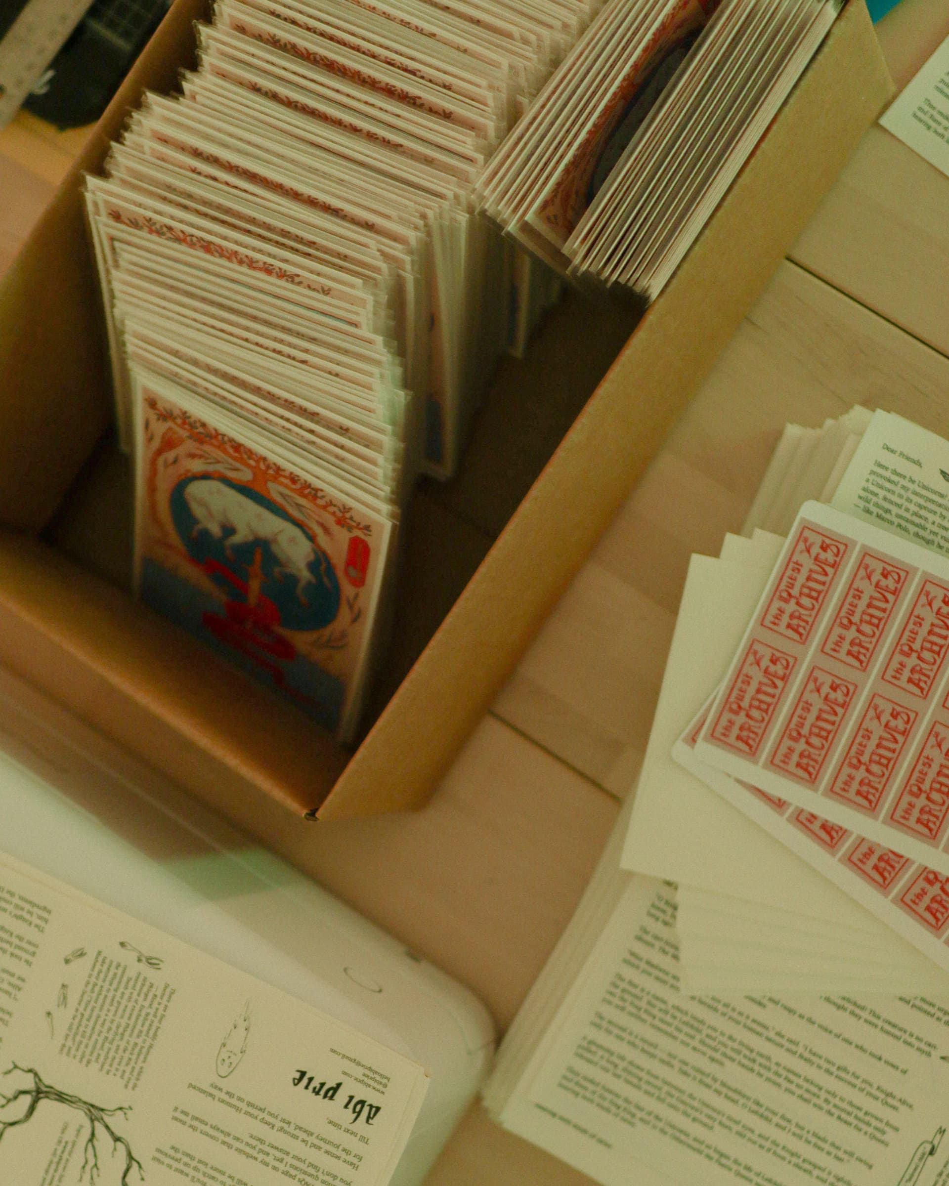 Cardboard box packed with sleeved Quest prints next to The Quest Archives stickers and letters