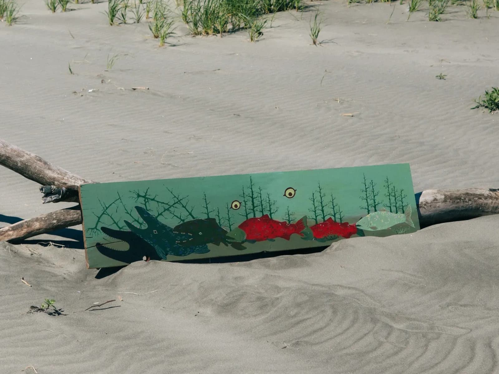 Image shows a painted plank in the sand. The plank is painted with salmon progressively turning into sea monsters, with trees growing on their backs.
