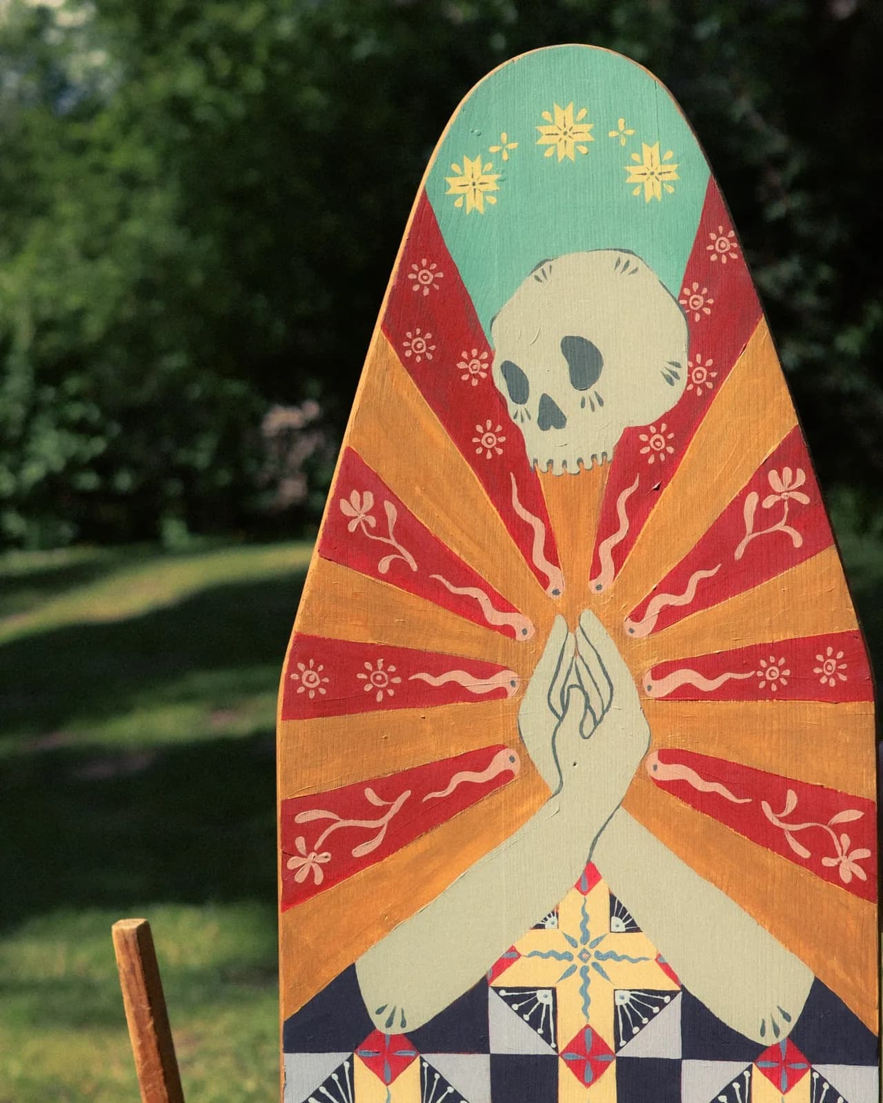 Image shows a woman in a blue shirt holding an ironing board. The ironing board is painted to look like a woman with a skull for a face, cloaked in a quilt of many colors. There are stars above her head, and a moth of death at the feet.